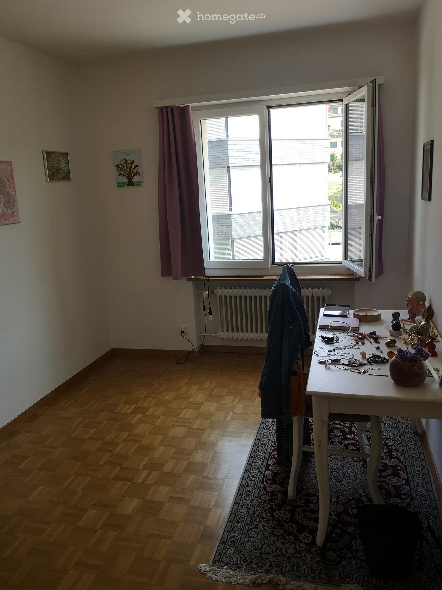 A small, well-lit room with wooden flooring, a large window with purple curtains, a white table with various items including a vase with flowers, and a radiator below the window.