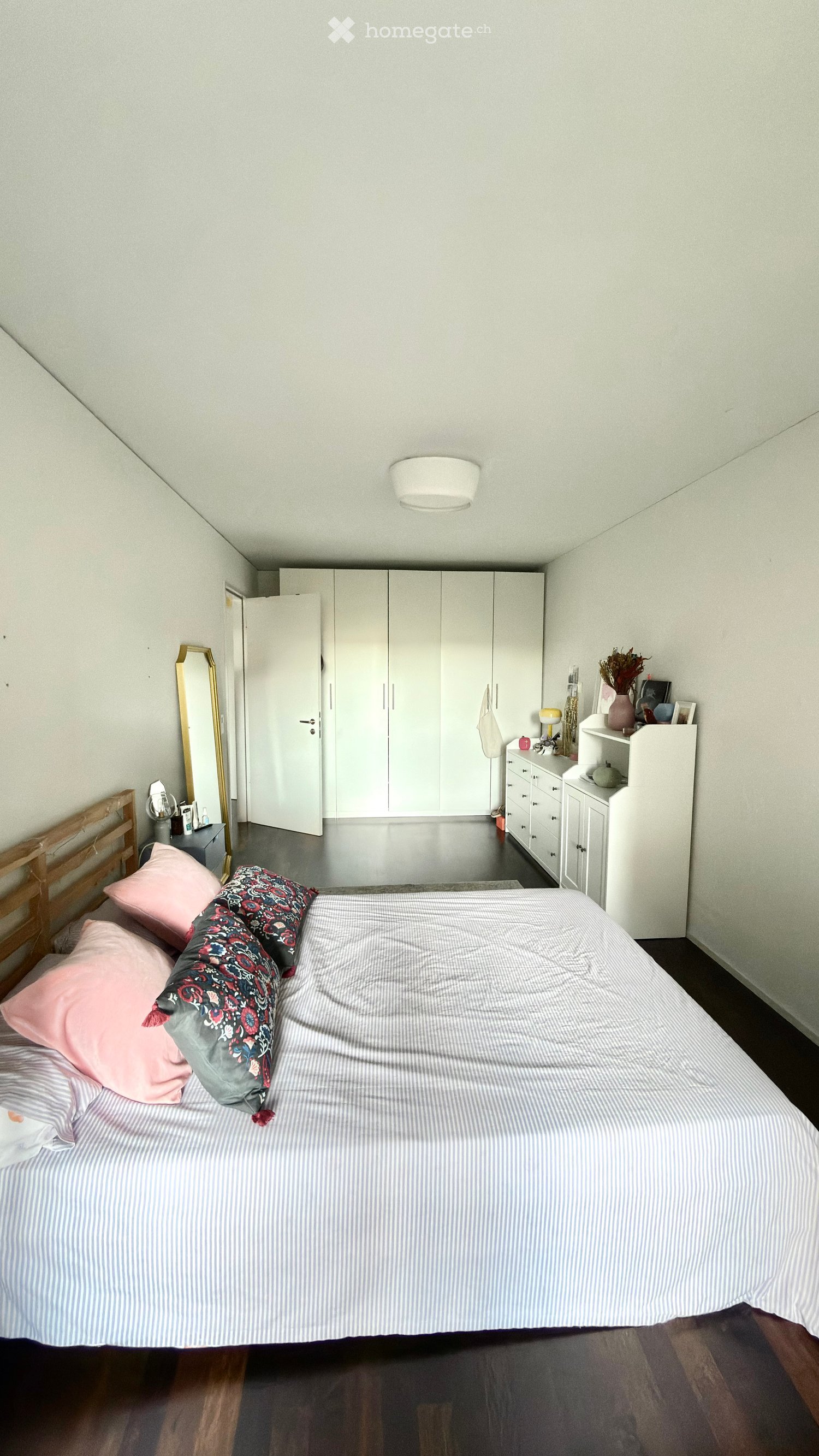A modern bedroom with a large bed, striped bedding, pink and patterned pillows, a wooden headboard, white wardrobe, a gold-framed mirror, and a nightstand with a lamp and decorative items.