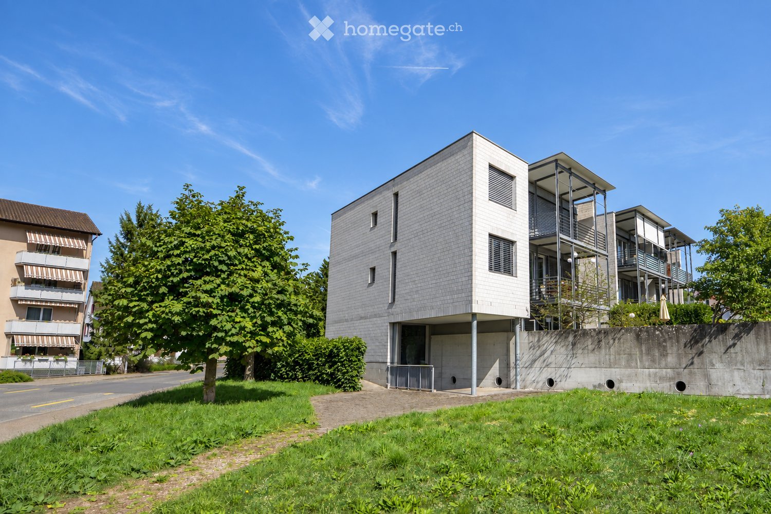 Modern, multi-story residential building with a raised ground floor, surrounded by greenery and a paved pathway.
