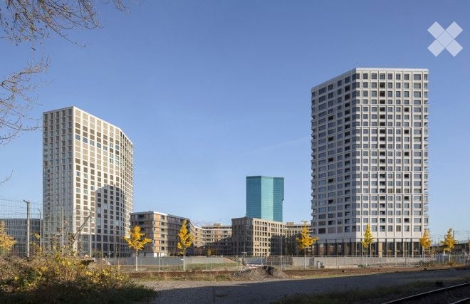 A modern urban development with multiple high-rise residential buildings and a central office tower, surrounded by trees and a fenced area.