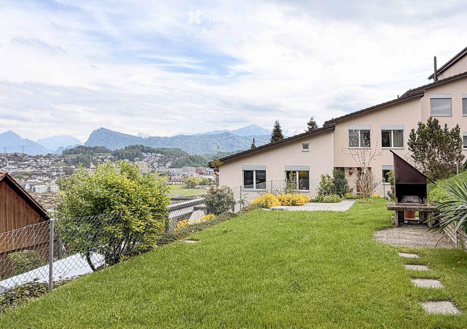 A two-story house with a well-maintained garden, featuring a barbecue area, a fence, and a scenic mountain view in the background.