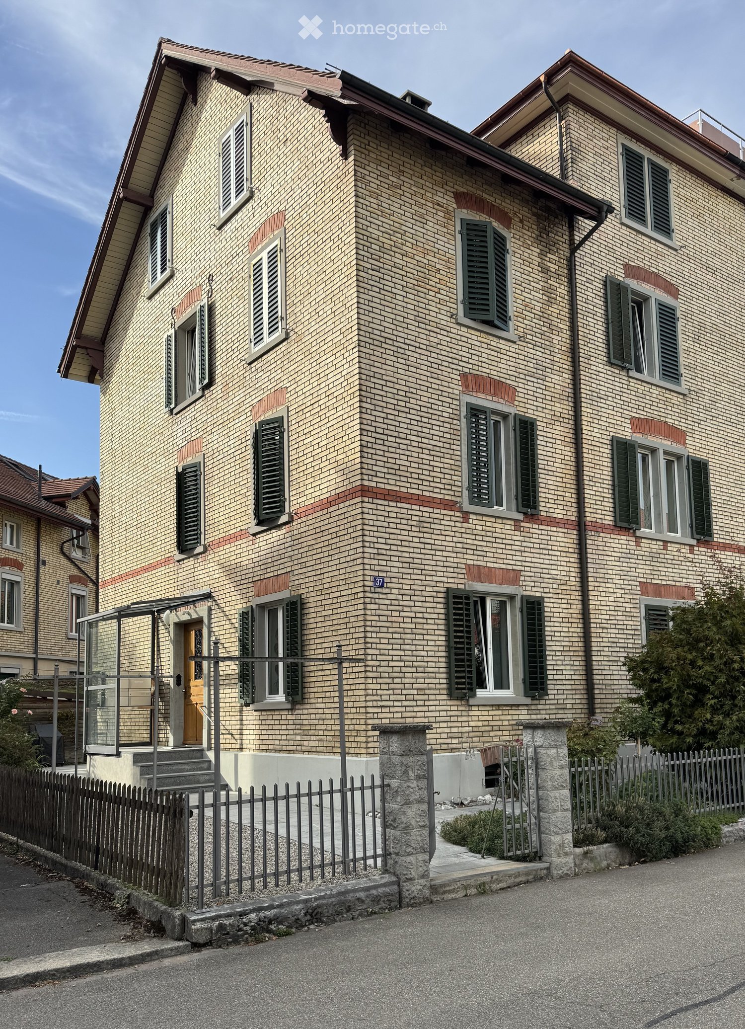 Two-story house with beige brick facade, green shutters, and a small balcony area with glass railing.
