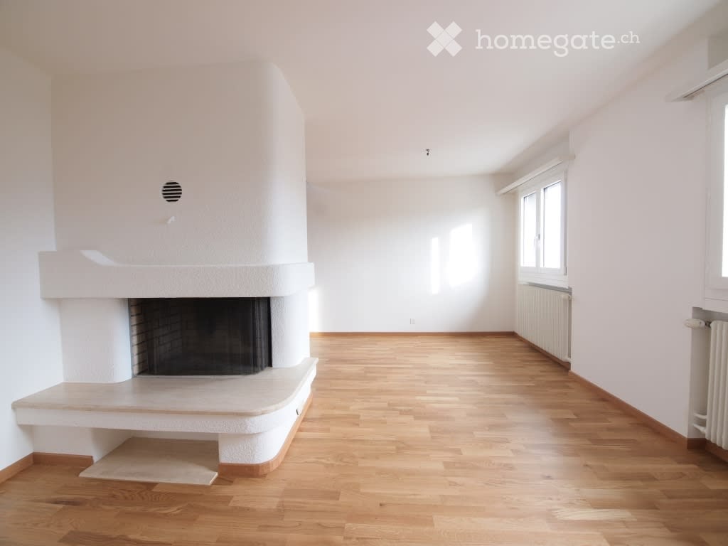 Empty room with light wooden flooring, white walls, and a built-in fireplace. Large windows allow natural light to fill the space.