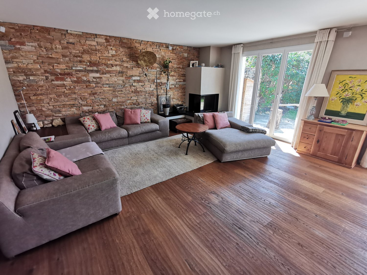 Modern living room with a stone accent wall, grey sectional sofa, coffee table, side tables, floor lamp, fireplace, and large windows with curtains.