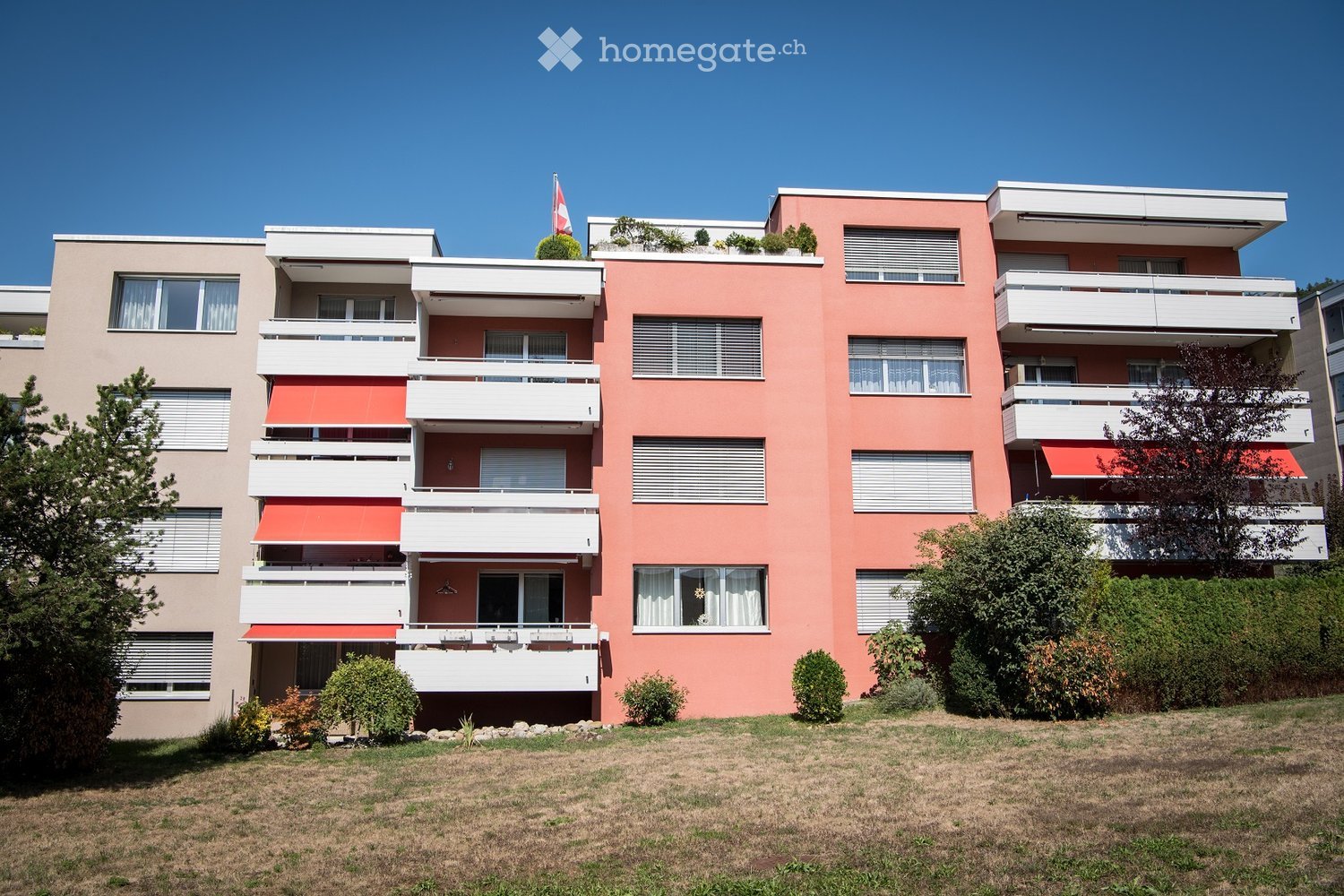A multi-story residential building with pink and beige facades, white balconies, and red awnings. The building features multiple windows with white frames and curtains, surrounded by greenery and a grassy lawn.