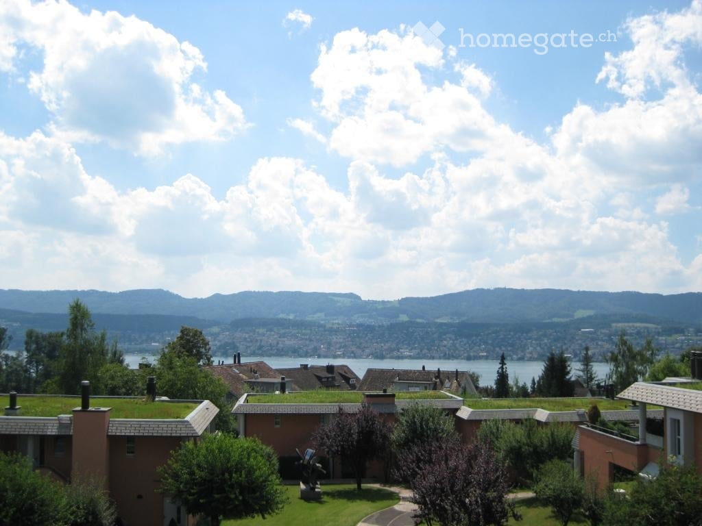 A modern building with a green roof, surrounded by trees and shrubs, overlooking a scenic lake and distant mountains.