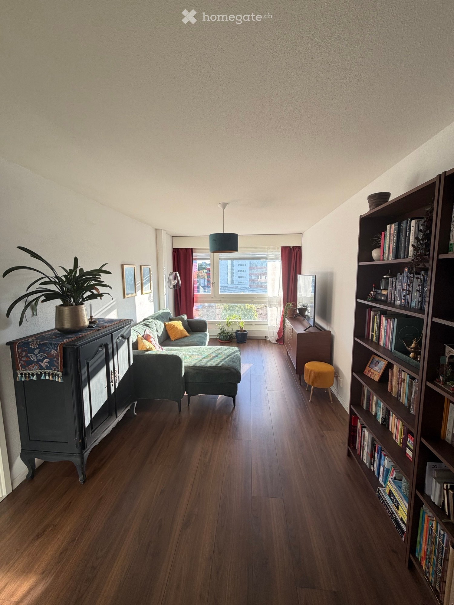 Living room with wooden flooring, a grey sofa with cushions, an ottoman, a bookshelf filled with books, a small yellow stool, a dark cabinet with a potted plant, and a mirror on a side table.