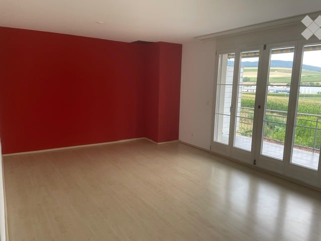 Empty room with wooden flooring, red accent wall, large glass sliding doors leading to a balcony with scenic countryside view.