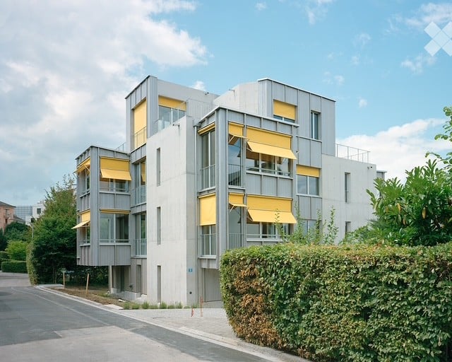 Modern multi-story building with grey facade, large windows, and yellow awnings. The building is surrounded by greenery and a neatly trimmed hedge.