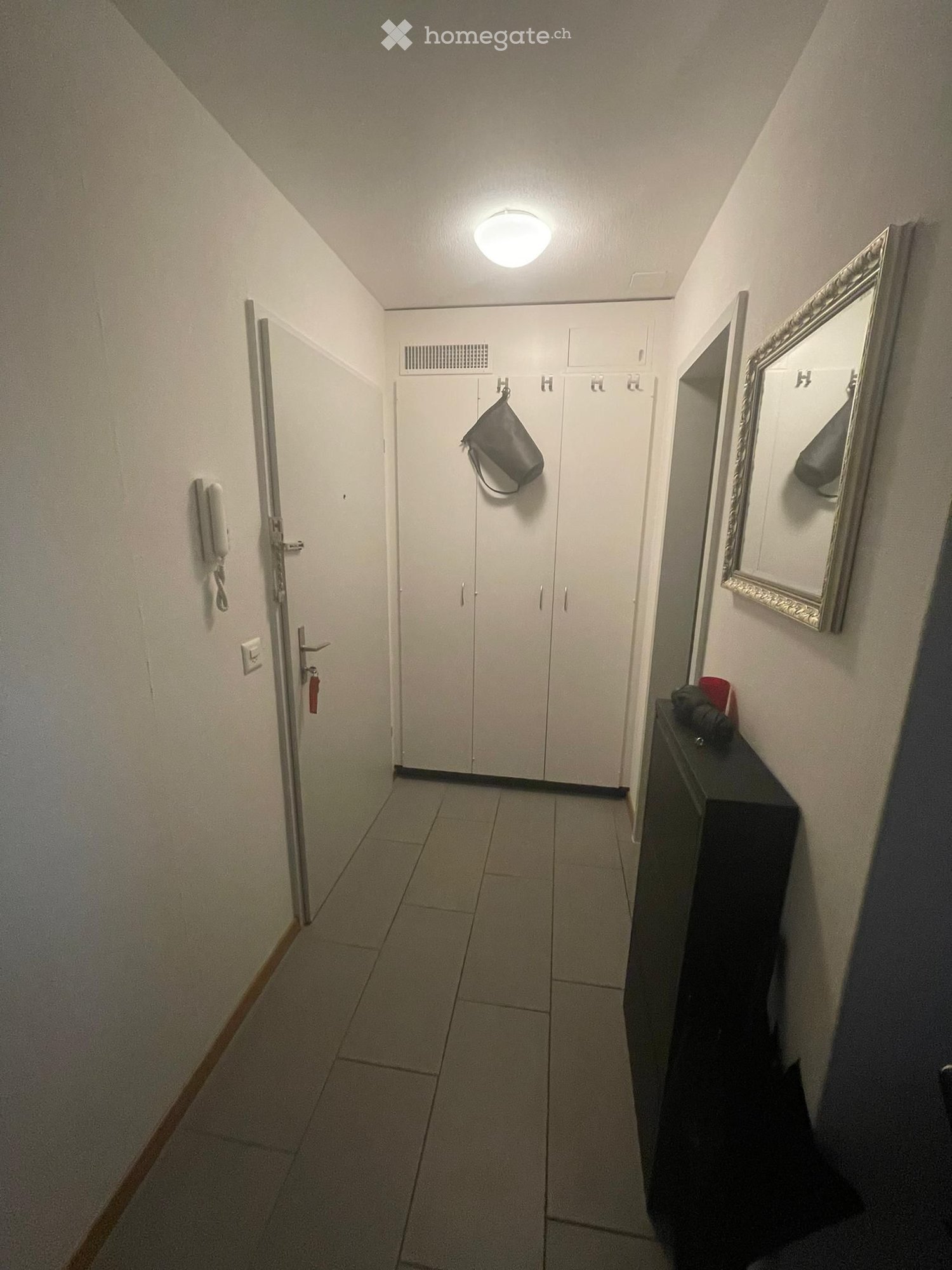 A narrow hallway with tiled flooring, white walls, and a ceiling light. Features include a closed door with a keycard reader, a small black table with a mirror, and a coat rack with a single bag hanging on it.