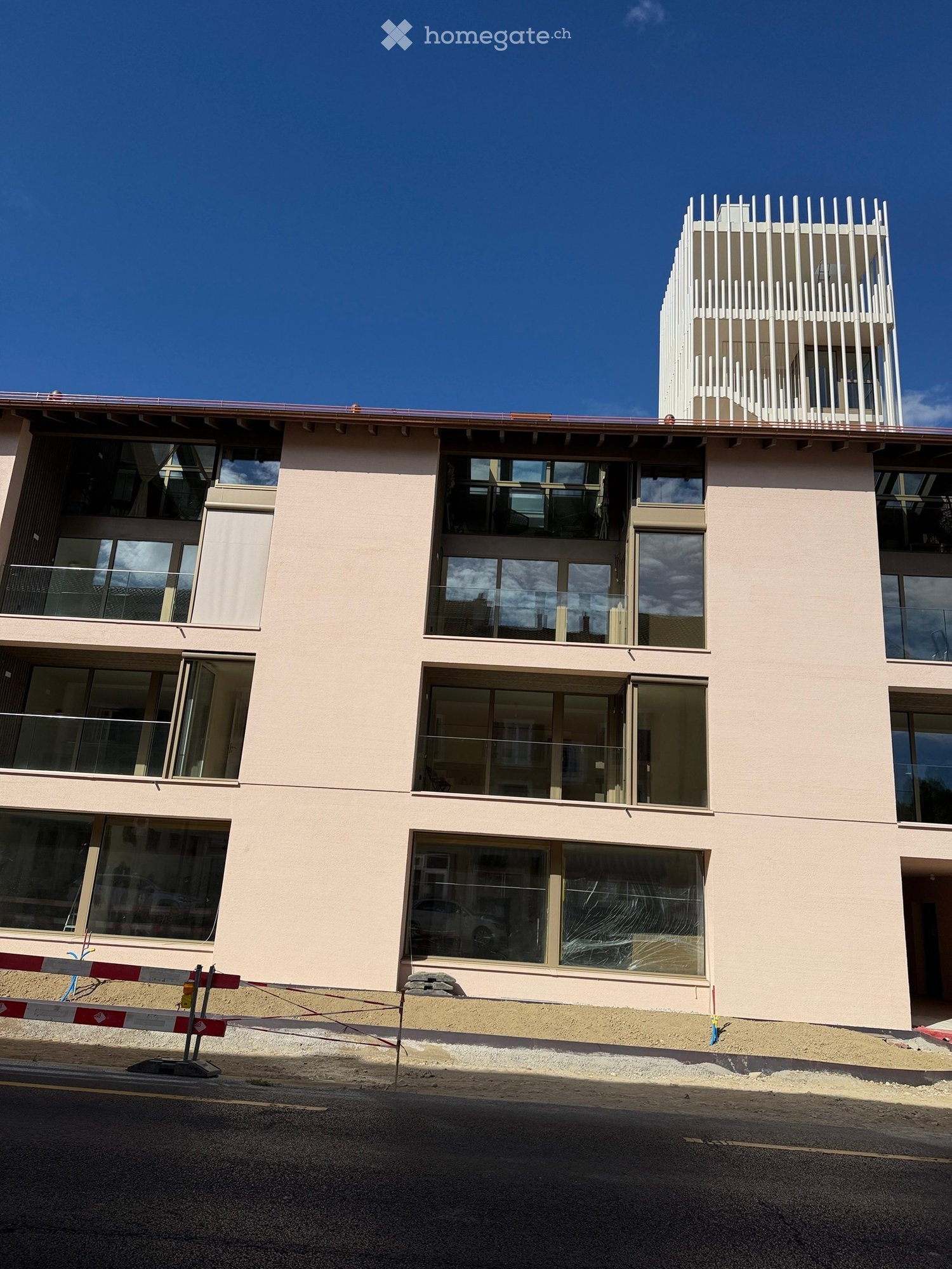 Modern multi-story building with large glass windows and balconies, light pink facade, and a contemporary design featuring white vertical slats on the roof.