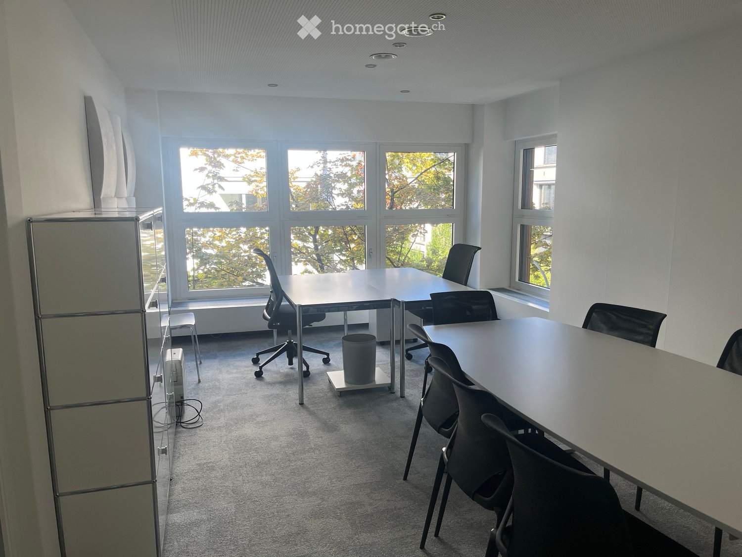 Bright, modern office space with large windows, multiple tables, chairs, and a cabinet.