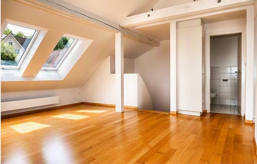 Bright, empty attic room with wooden flooring, white walls, skylight windows, and a small adjacent bathroom.