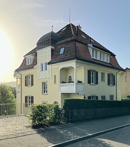 A two-story yellow house with white trim, multiple windows, a balcony on the first floor, and a well-maintained garden in front.