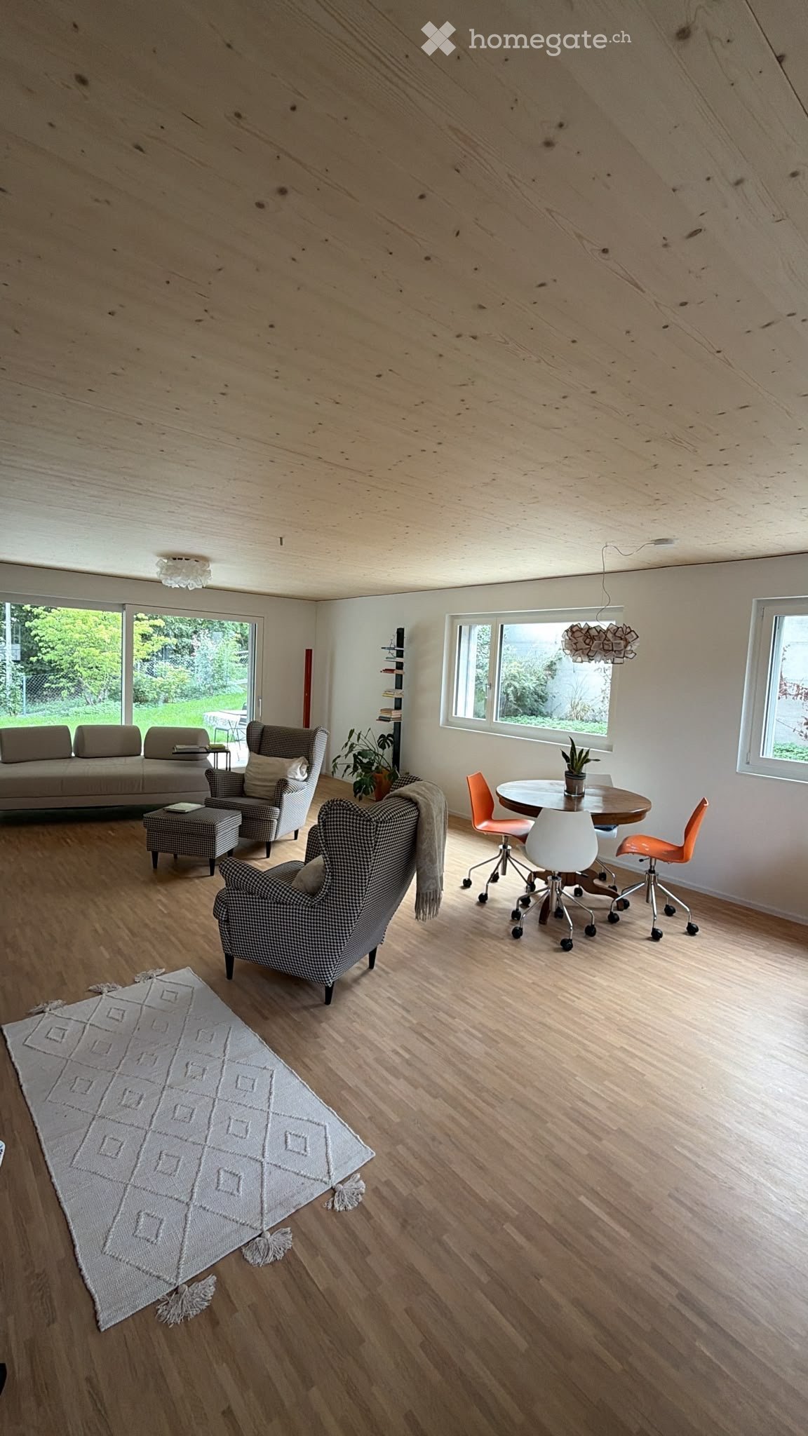 Open-plan living and dining area with wooden flooring, large windows, a sofa, armchairs, ottoman, dining table, chairs, and a rug.