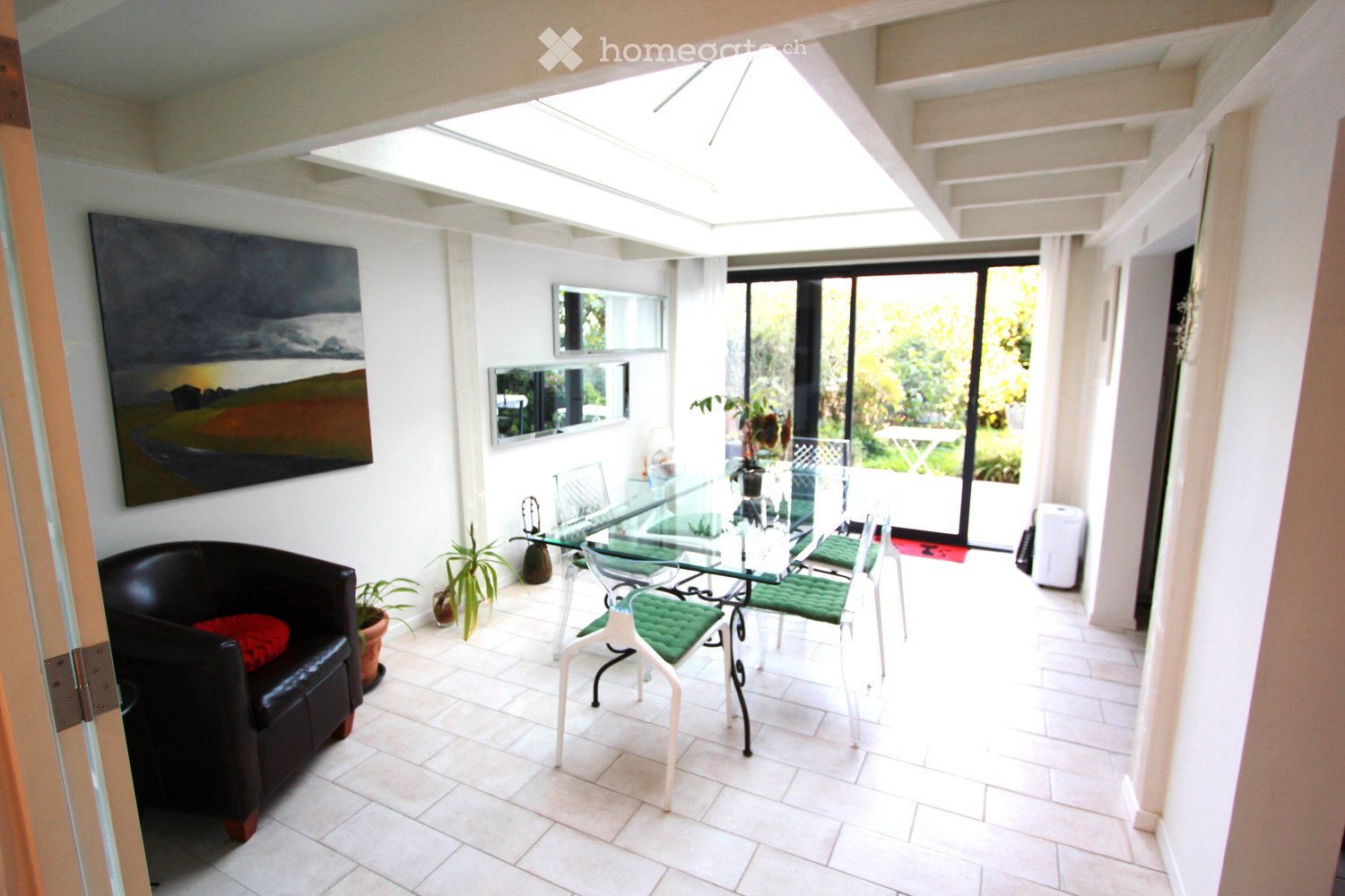 Bright, modern living and dining area with white tiled flooring, a black leather sofa, a glass dining table with green upholstered chairs, and a large painting on the wall. The space features large windows and a sliding glass door leading to an outdoor ar