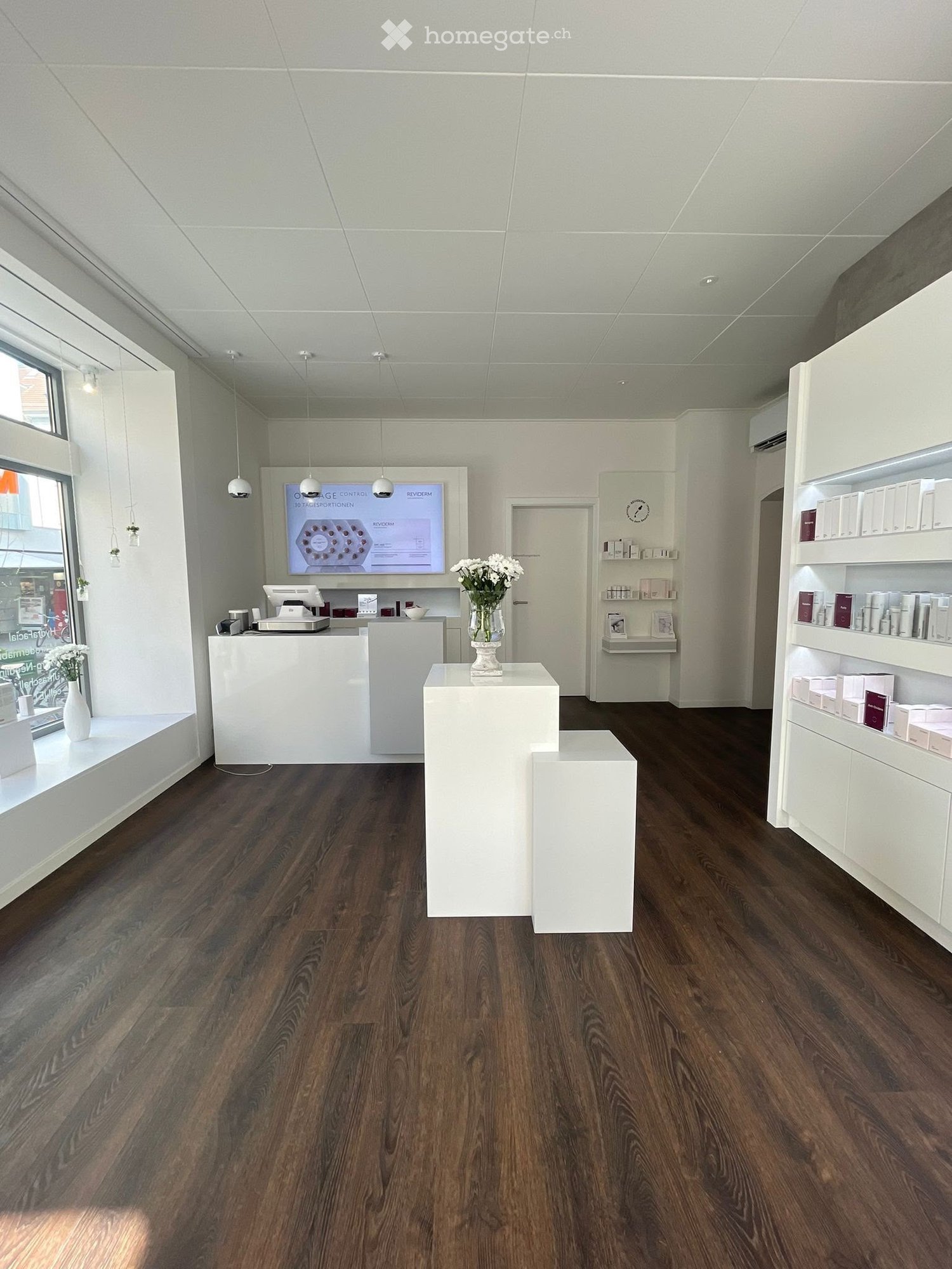 Modern, minimalist reception area with a polished wooden floor, white reception desk, large screen, decorative vase with flowers, and shelves displaying various products.