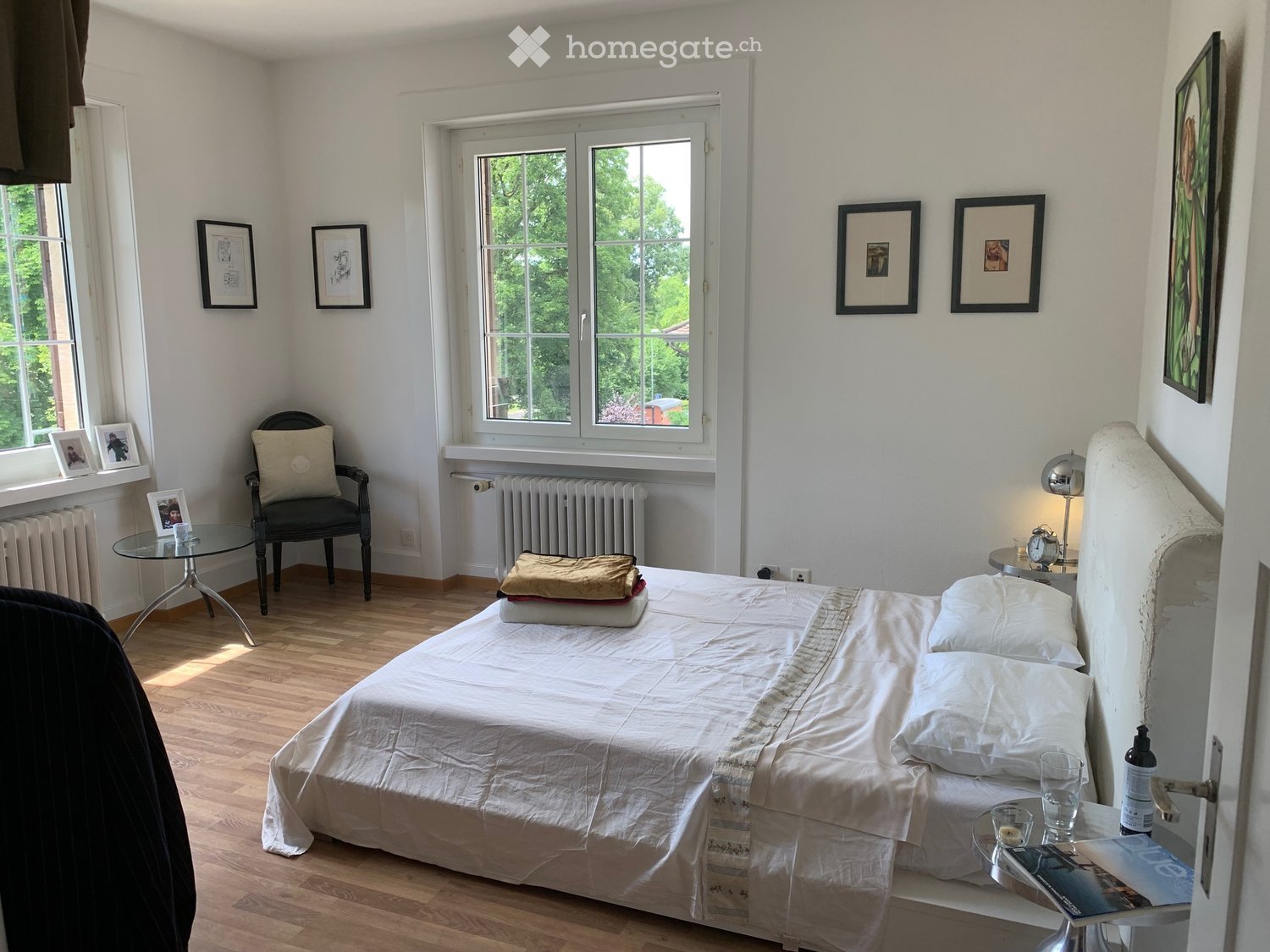 Cozy bedroom with wooden flooring, large windows, a neatly made bed, a side table with a lamp, a small round table, and a chair with a cushion.