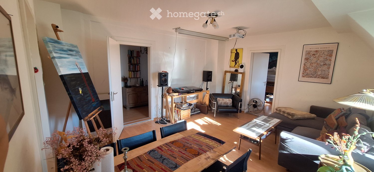 A well-lit living room with a wooden floor, a sofa, a dining table with chairs, a lamp, a fan, speakers, a mirror, a cabinet, and a painting on an easel.