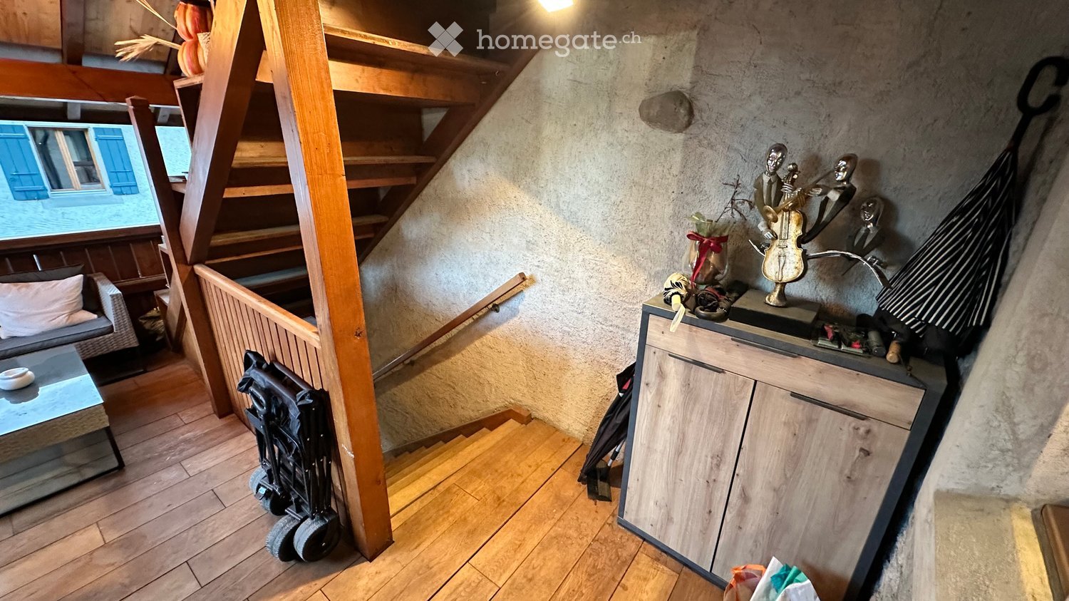 A cozy, well-lit room with wooden flooring, a staircase leading upwards, a sofa with a coffee table, a stroller, a cabinet with decorative items including a statue and a lamp, and a hammock hanging on the wall.