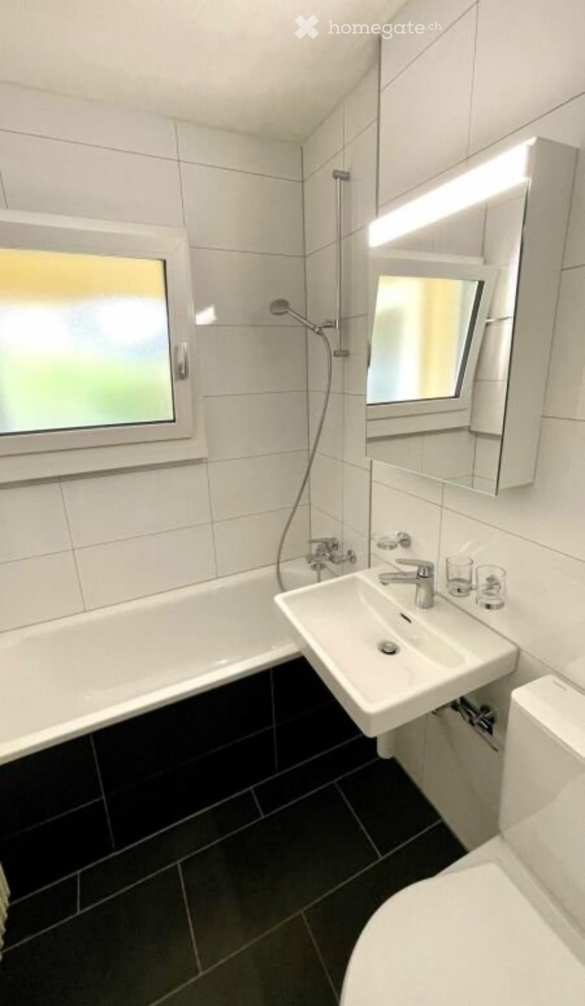 Modern bathroom with white tiles, black floor, bathtub, sink, toilet, and mirror.