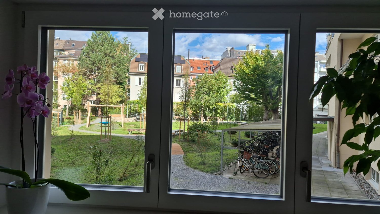 View from a balcony showing a green park area with trees, playground equipment, and a paved path with bicycles parked nearby.