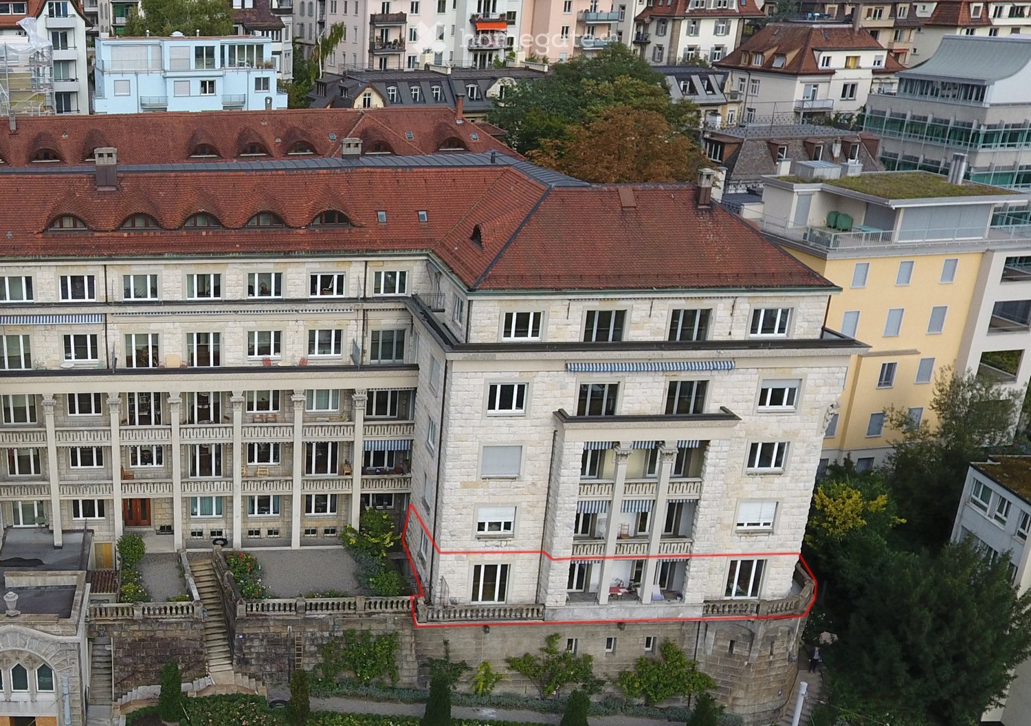 A multi-story residential building with a red-tiled roof, white stone facade, and multiple windows and balconies. The building is situated in a dense urban area with neighboring structures visible in the background.