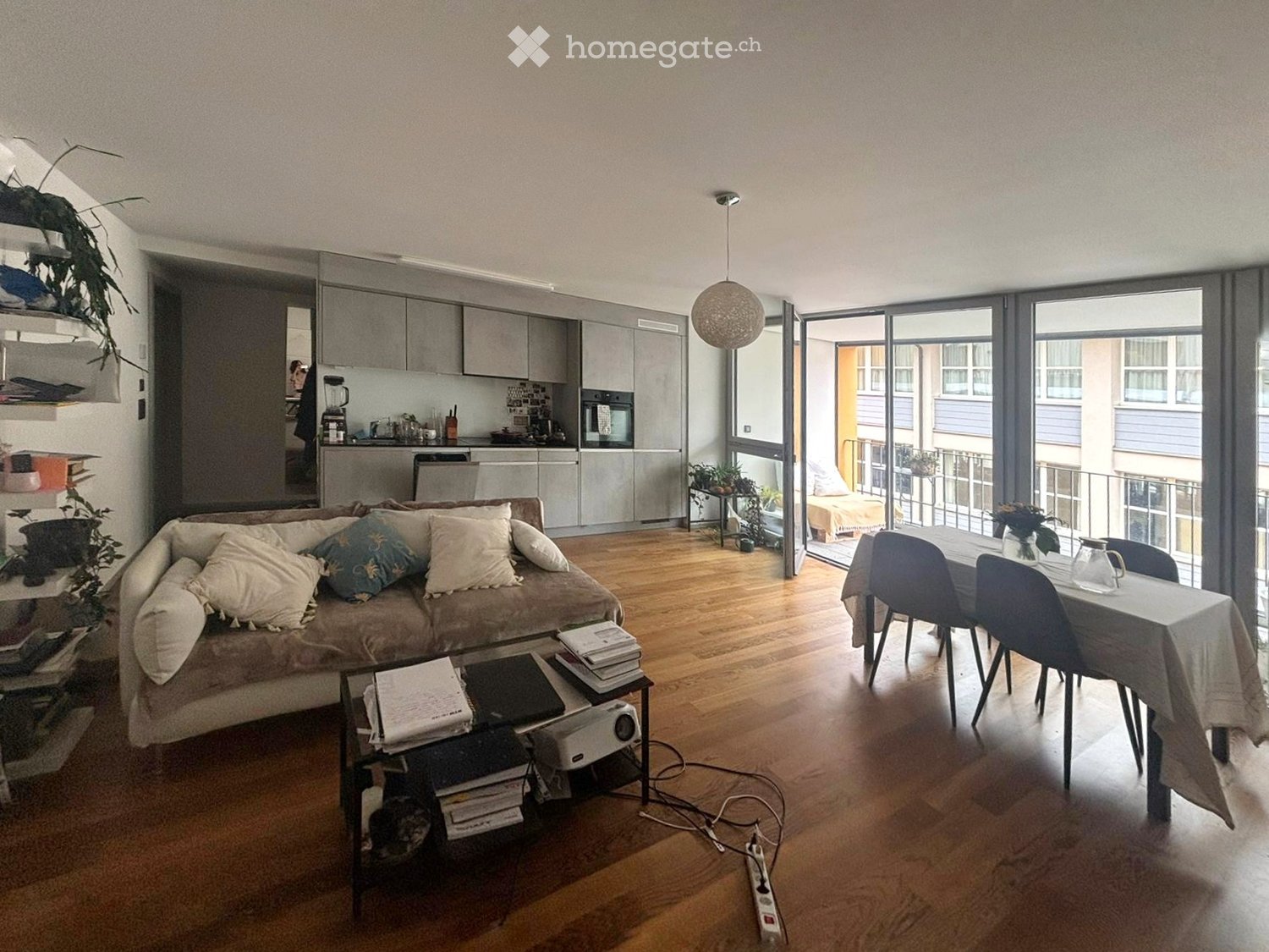 modern open living space, kitchen with modern appliances, large windows with terrace, dining table and chairs, couch with pillows, coffee table, desk, projector