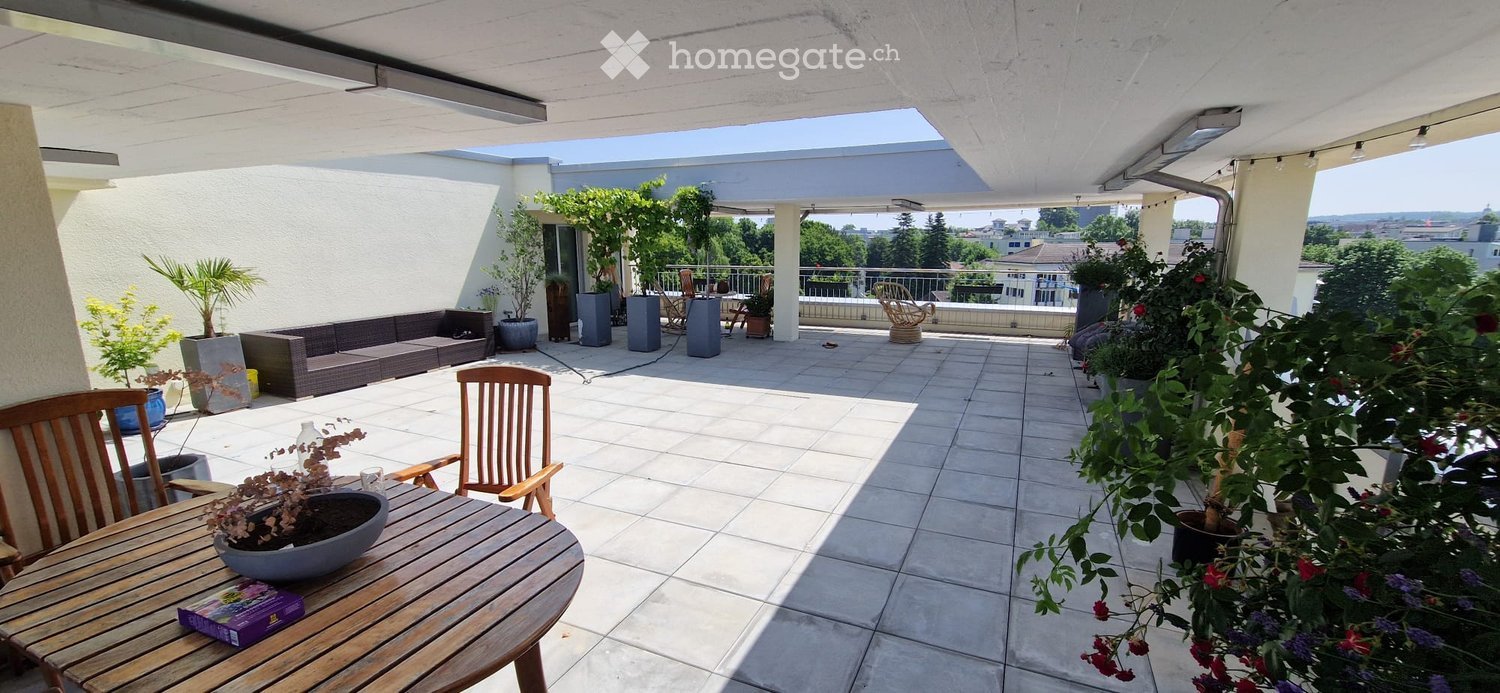 Spacious balcony with tiled floor, wooden table and chairs, sofa, potted plants, and scenic city view.