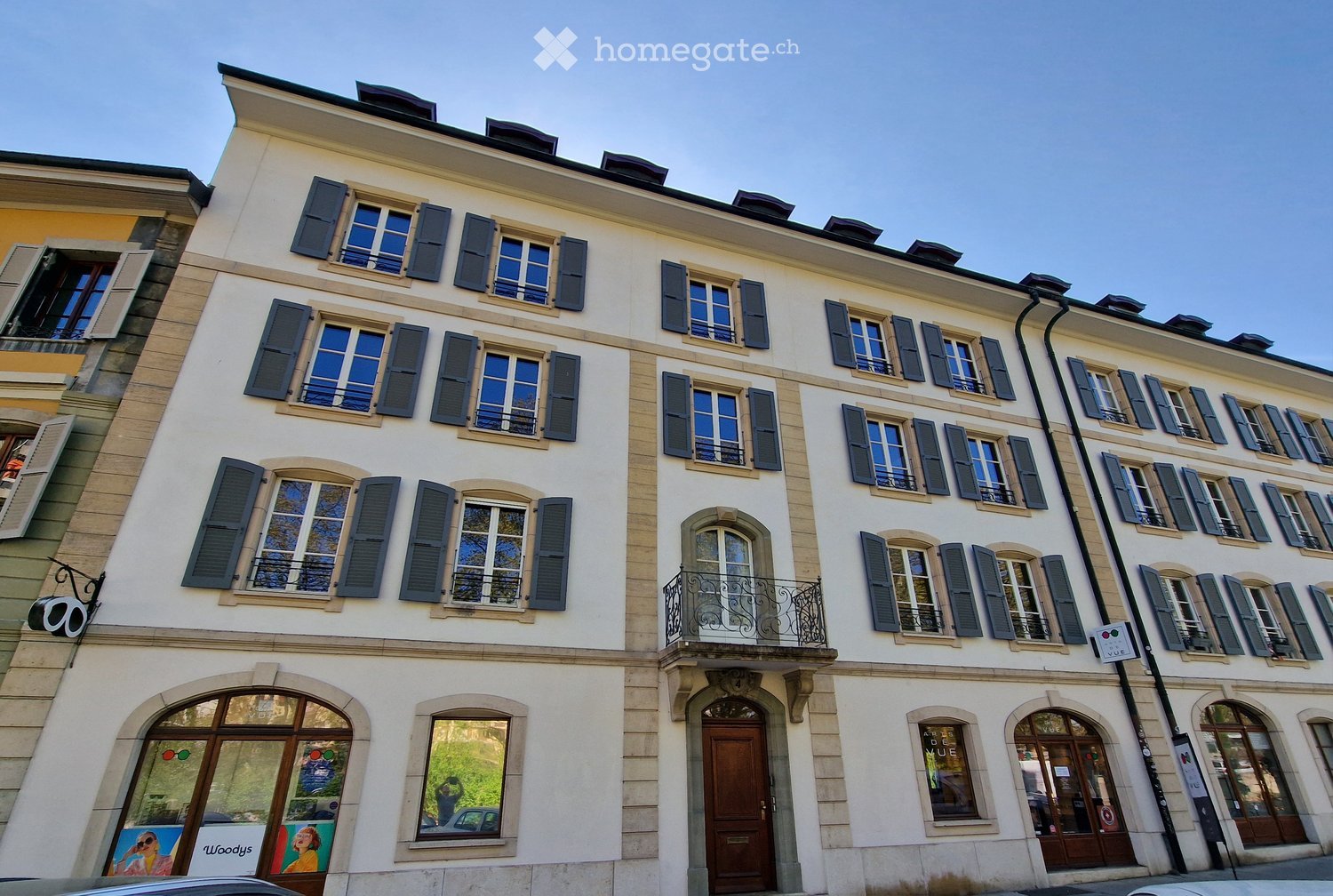 A multi-story residential building with white walls, dark shutters, and multiple windows. Features include a balcony with a metal railing, arched doorways, and a storefront with glass windows and signage.