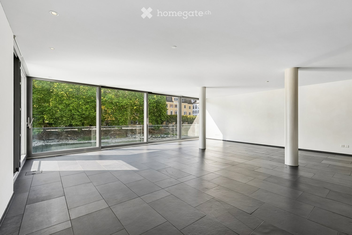 Empty room with dark grey tiled floor, white walls, large glass sliding doors leading to a balcony, and two white columns.