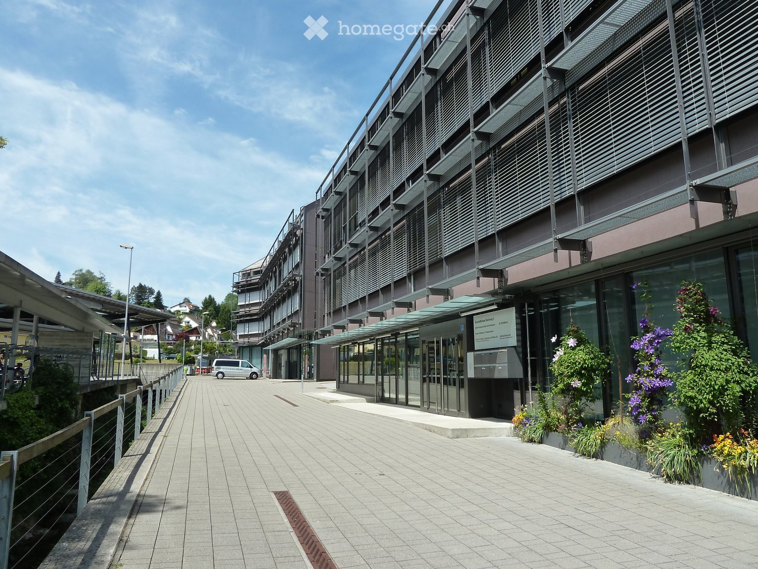 Modern multi-story building with glass facade, green shutters, and a paved walkway leading to the entrance.