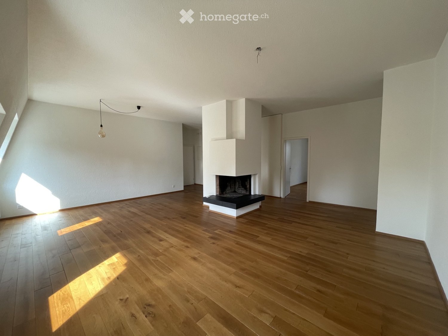 Empty room with wooden flooring, white walls, and a fireplace. Natural light streams in from a window on the left side.