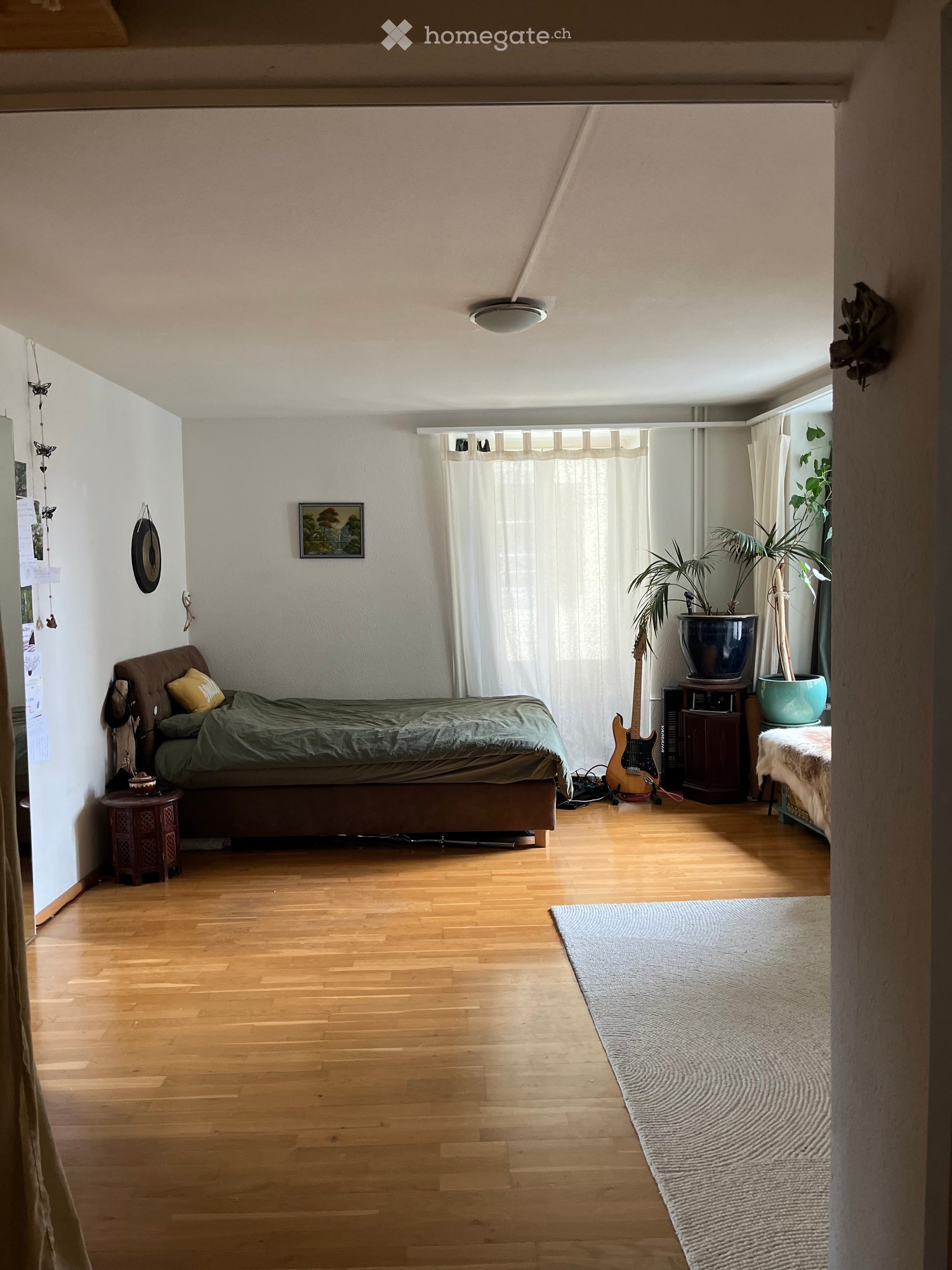 Cozy bedroom with wooden flooring, a bed with green bedding, a guitar leaning against the wall, a nightstand, a small table with a plant, and a large window with white curtains.