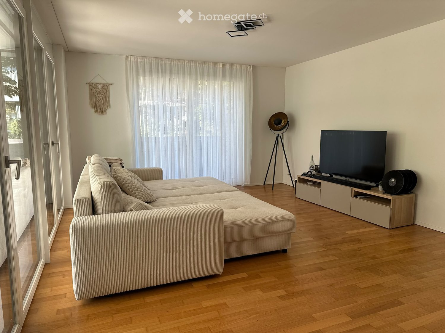 Modern living room with a large sectional sofa, coffee table, flat-screen TV on a wooden stand, floor lamp, and sheer curtains covering large windows.