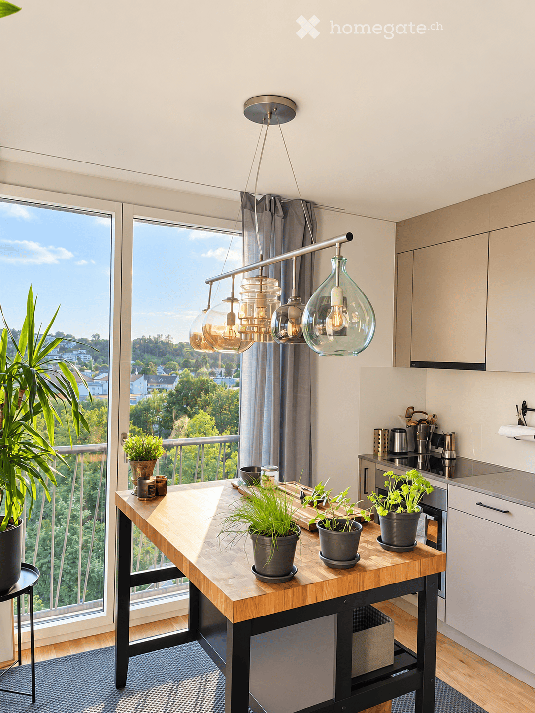 Modern kitchen with a wooden island, pendant lights, potted plants, and a view of greenery outside through large windows.