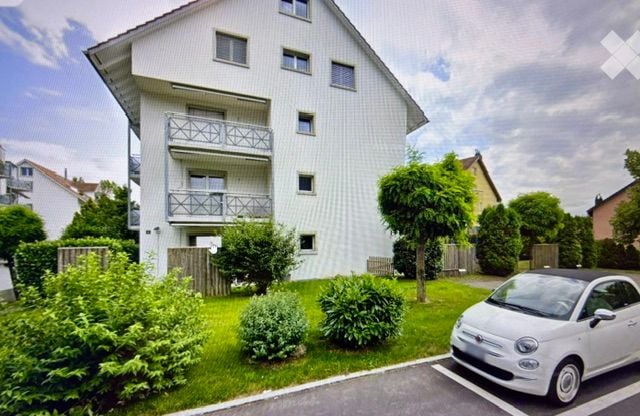 Modern multi-story apartment building with white facade, multiple balconies, and surrounding greenery. A small white car is parked in the driveway.