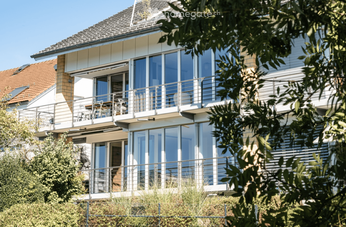Modern two-story building with large windows, balconies, and a tiled roof, surrounded by greenery.