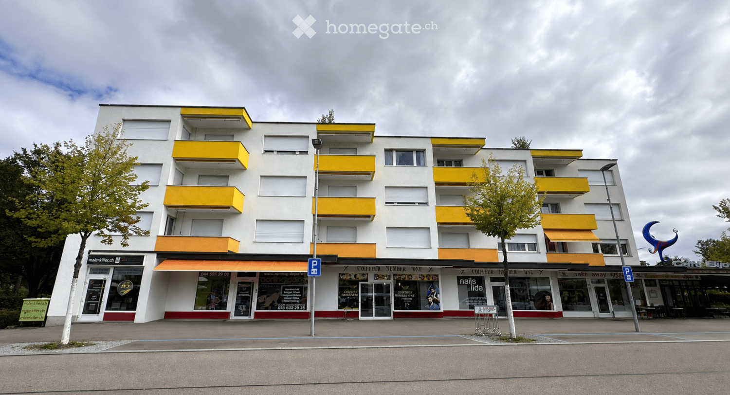 Modern multi-story building with white facade and yellow balconies, ground floor commercial spaces with storefronts and signage.