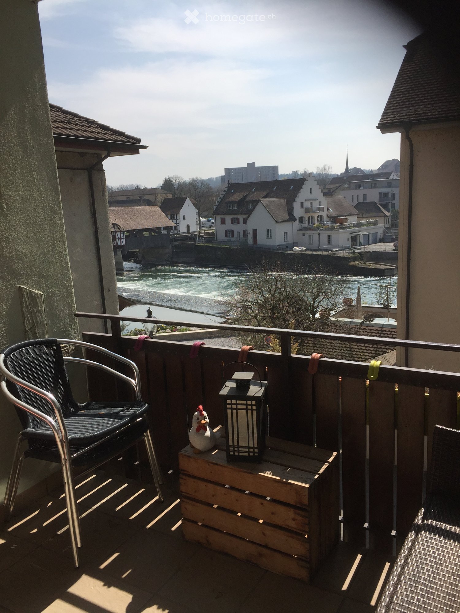 Balcony with a view of a river and town, featuring a wooden table, lantern, figurine, and two chairs.