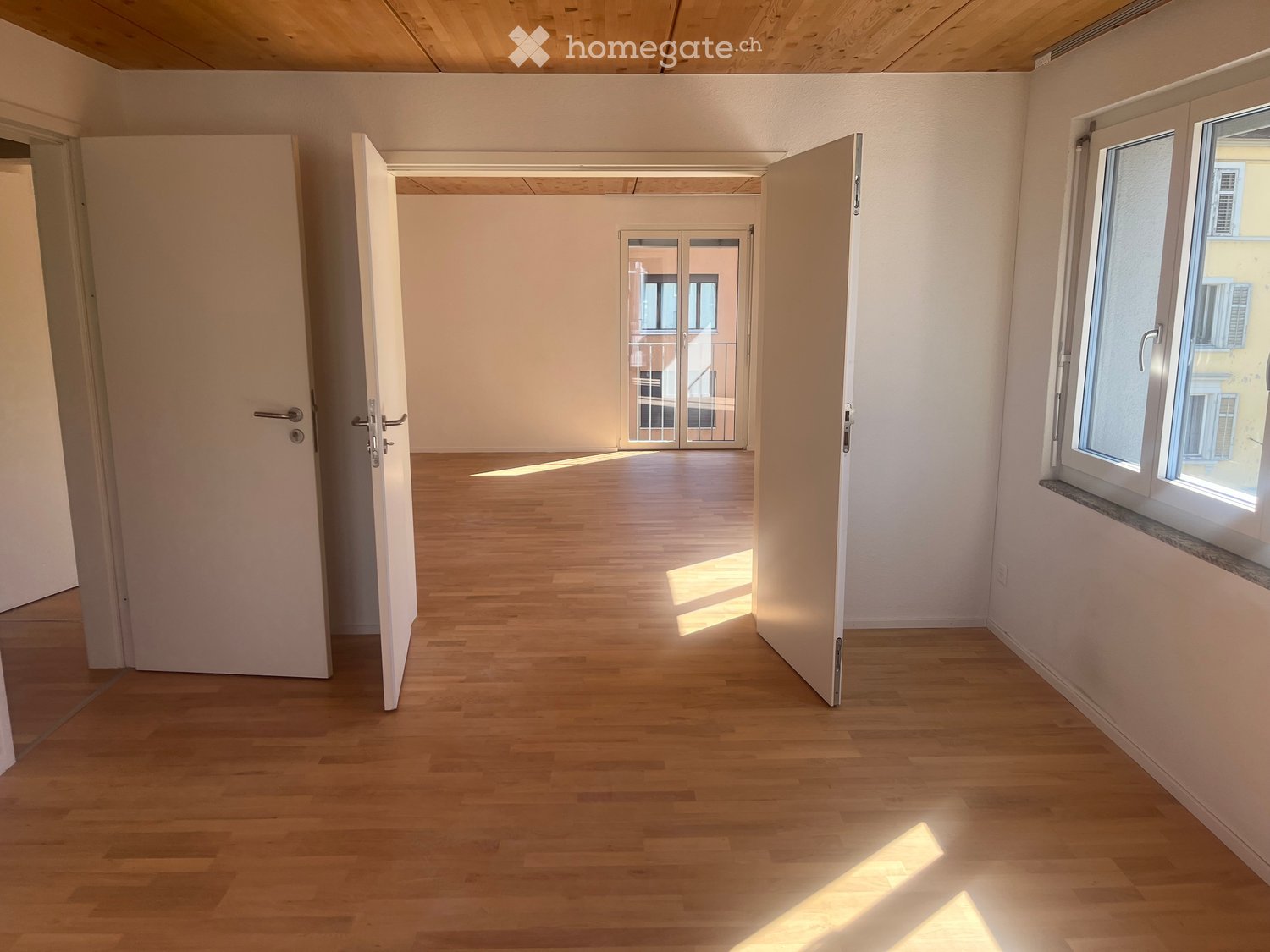 Empty room with wooden flooring, white walls, and large windows allowing natural light. The room features an open doorway leading to another space and a sliding glass door to a balcony.
