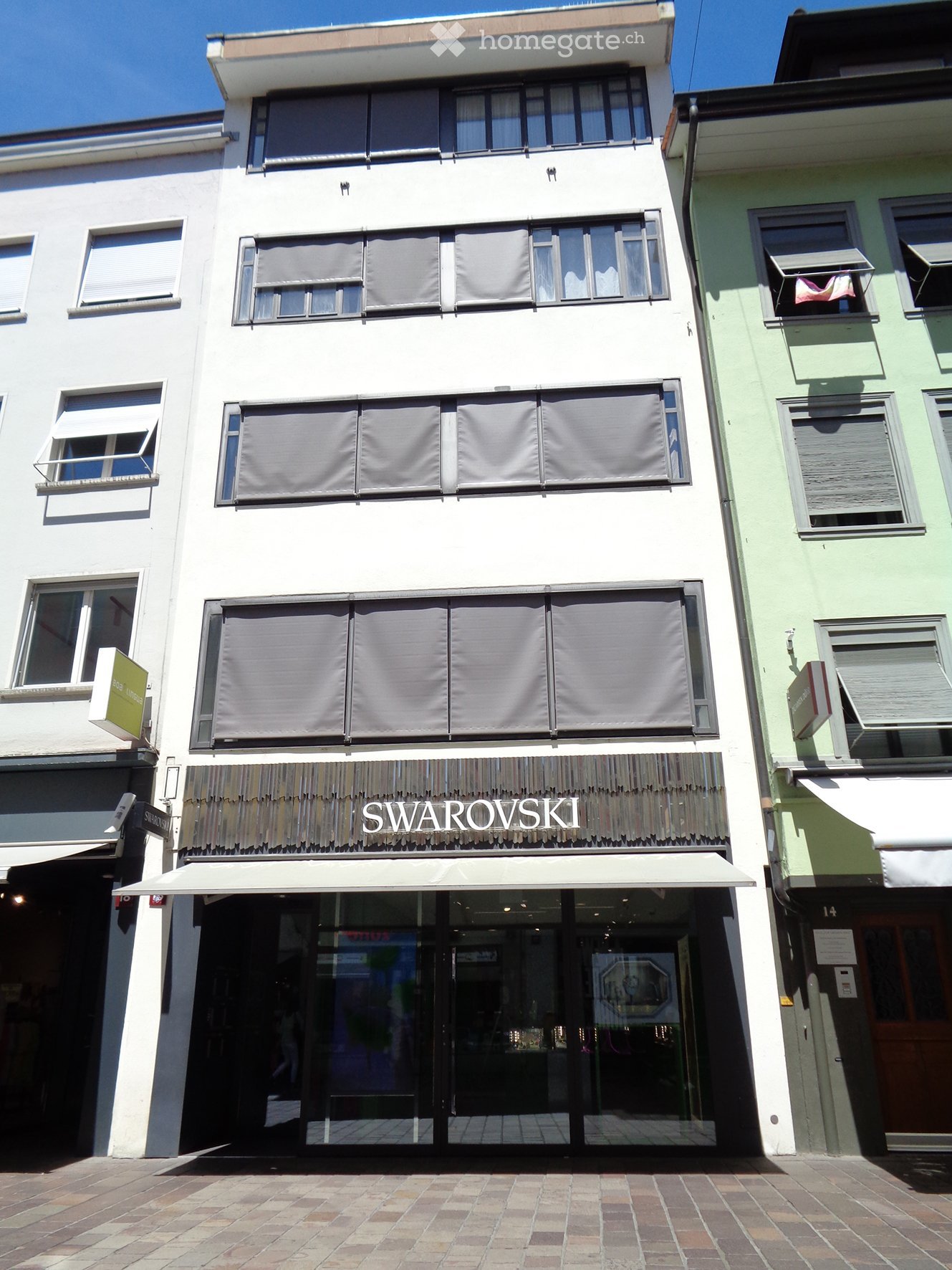 A three-story building with a storefront labeled "SWAROVSKI" and a white awning over the entrance. The facade is white with multiple windows, some covered by blinds.