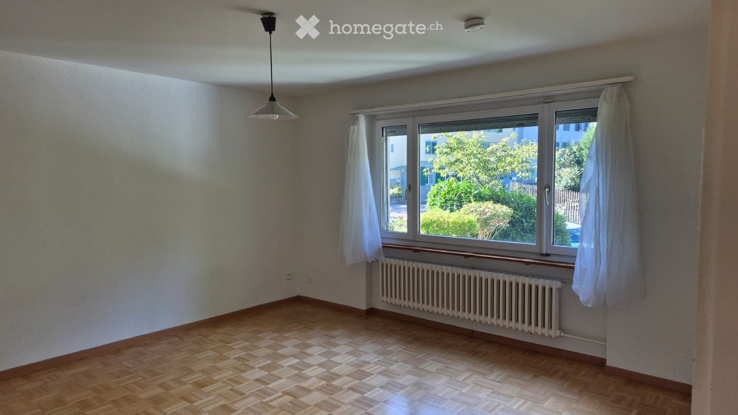 Empty room with wooden flooring, large windows with white curtains, radiator, and a ceiling light fixture.