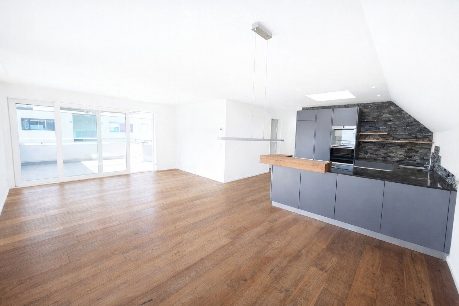 Modern kitchen with grey cabinets, wooden countertop, and black stone backsplash. Features a large open space with wooden flooring and large windows.