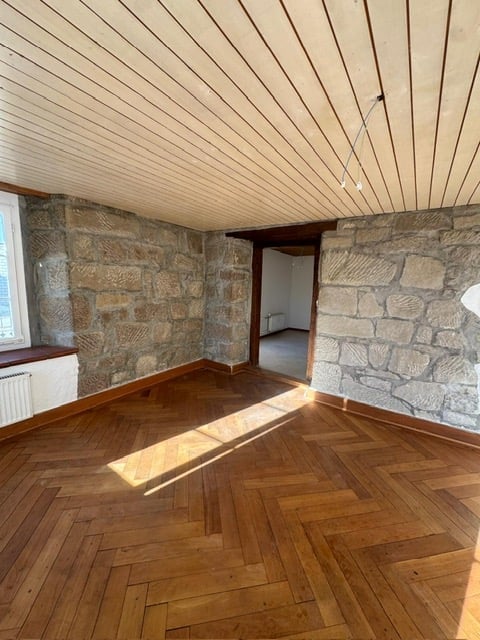 Empty room with exposed stone walls, herringbone wooden flooring, and a slanted wooden ceiling. Natural light streams in from a window on the left.