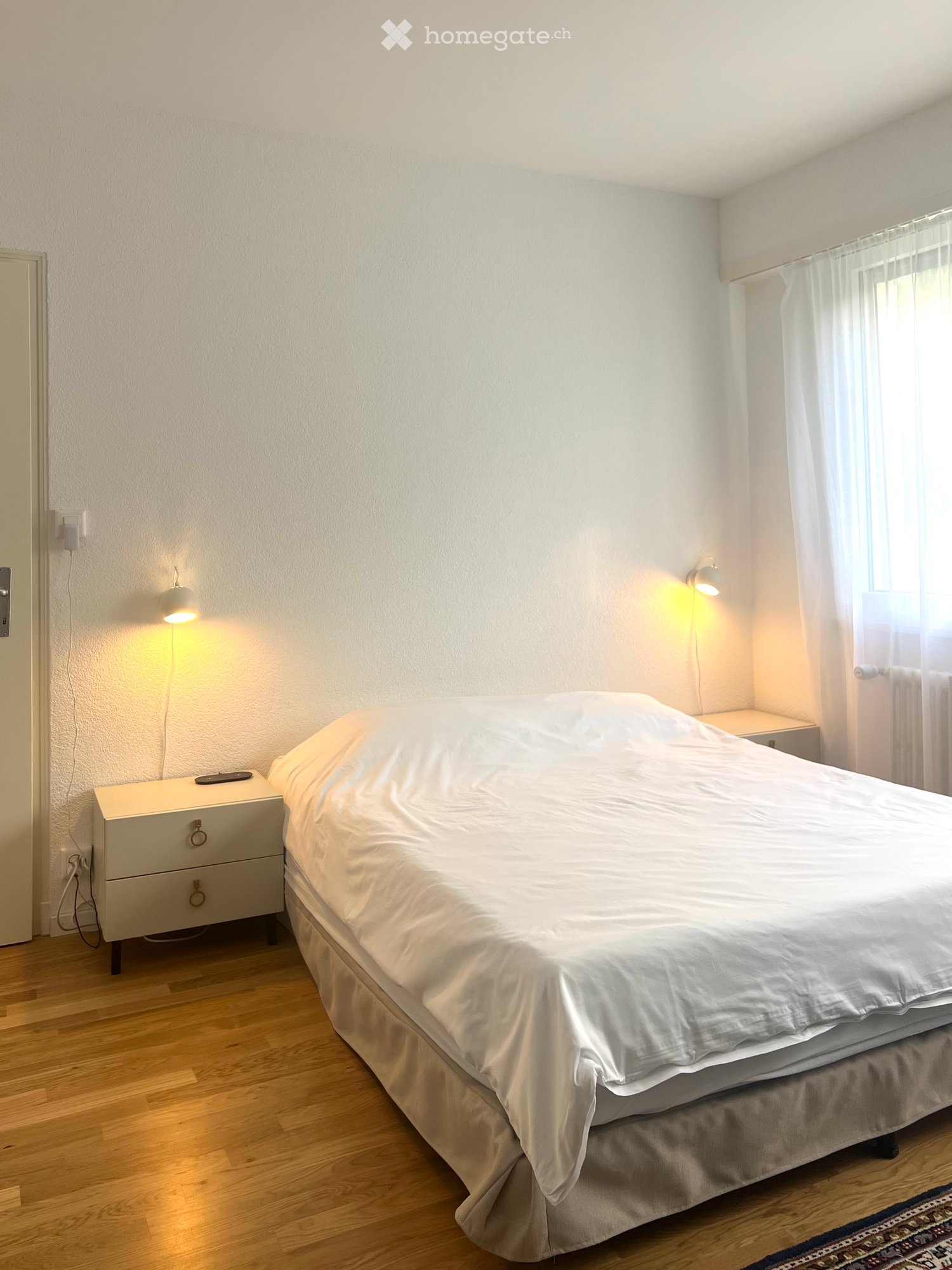 A neatly arranged bedroom with a large bed covered in white bedding, a nightstand with a lamp, and a wooden floor.