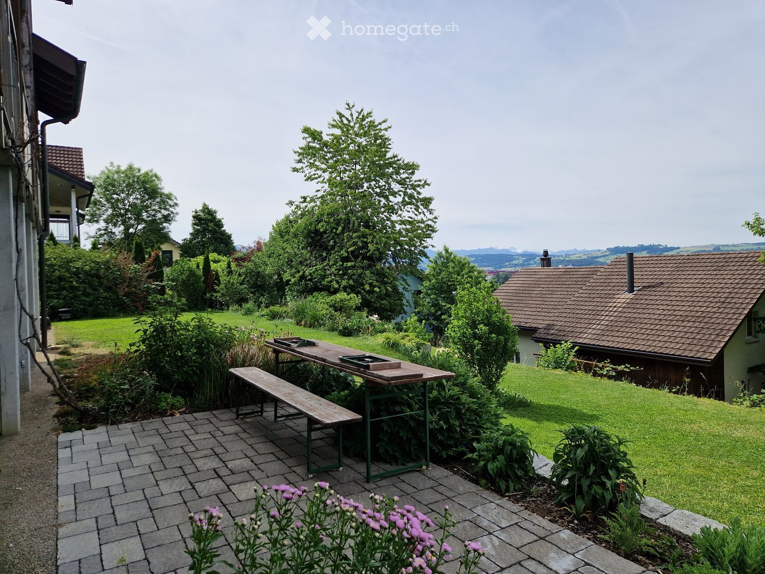 Garden area with a wooden table, bench, and surrounding greenery, including flowers and trees, with a view of a house and landscape in the background.