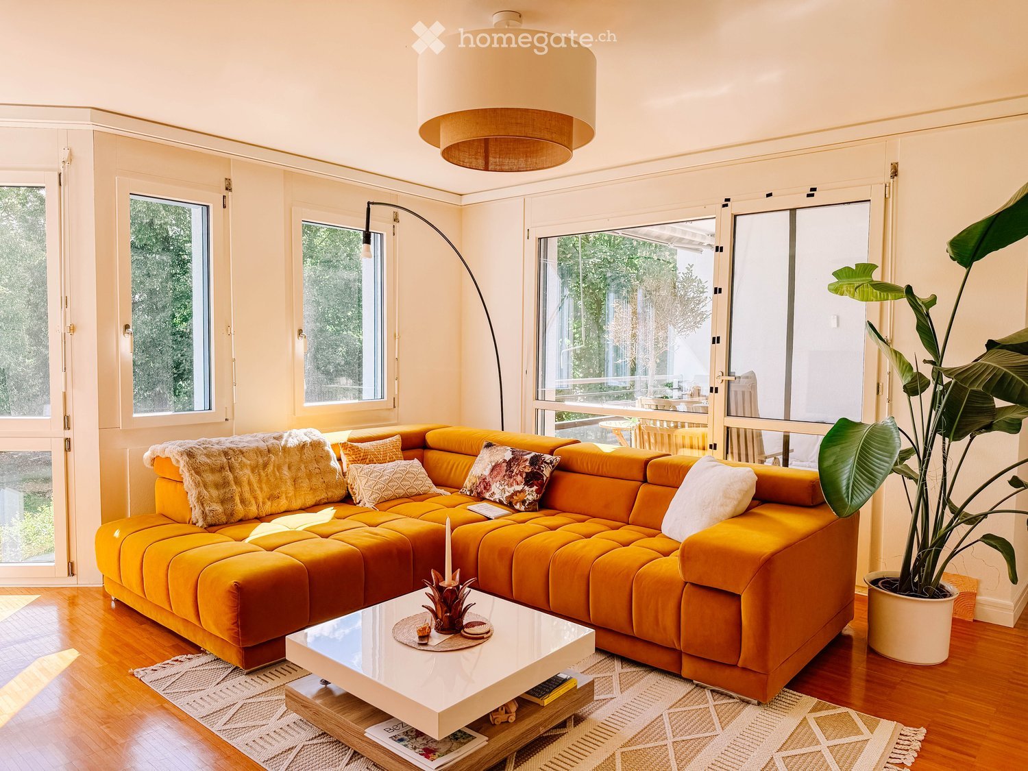 Bright living room with large windows and sliding glass doors leading to a patio. Features a mustard yellow sectional sofa with throw pillows and a fur throw, a white coffee table with a candle and decorative items, a floor lamp, and a large potted plant.