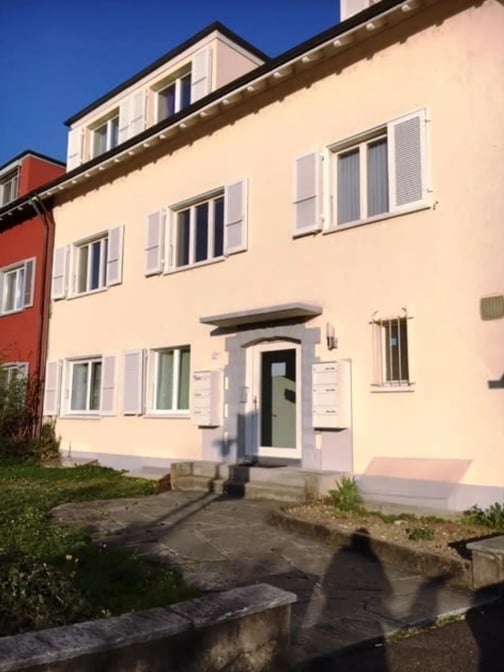 A three-story residential building with light pink facade, white shutters, and multiple windows. The entrance features a small porch with steps leading up to a green door.