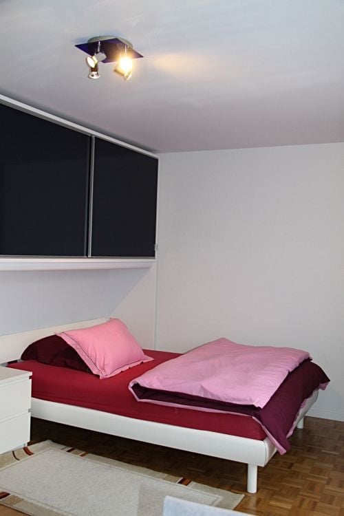 A modern bedroom with a white bed frame, red bedding, pink pillows, and a matching pink blanket. The room features wooden flooring, white walls, and built-in dark-colored cabinets above the bed.